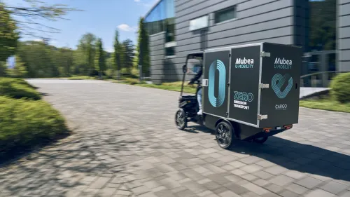 E-Cargobike mit großem Laderaum fährt auf gepflastertem Weg an modernen Gebäude entlang.