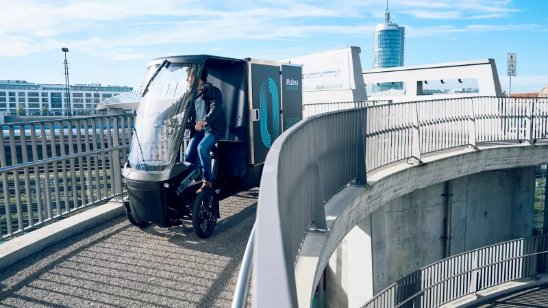 Cargo PACK auf einer Fahrradbrücke in München vor einer Skyline moderner Gebäude.